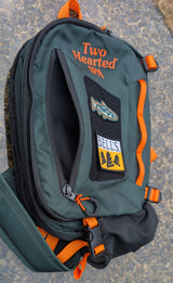 A dark green and black Two Hearted IPA backpack with bright orange accents, featuring a side pocket with a fish patch and a BELLS logo patch.