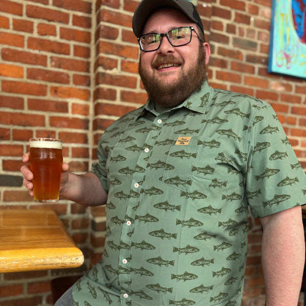 green short sleeve button up collar shirt with an allover print of dark green trout. there is a beige pinch tag on the only pocket with Two Hearted IPA stitched into it.