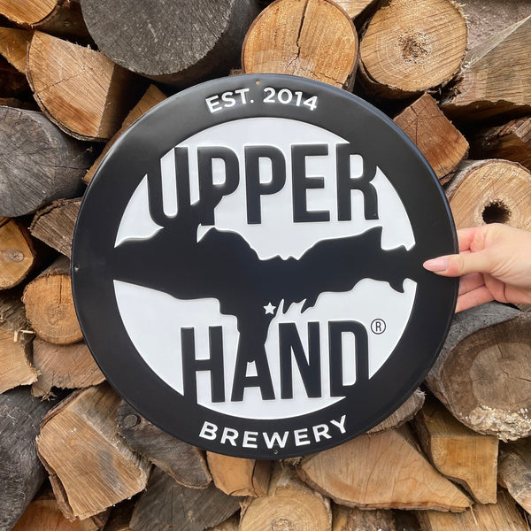 Hand holding a round tin sign of the Upperhand logo. The background is a stack of chopped wood.