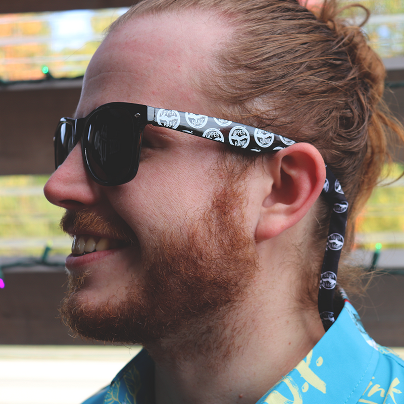 Bearded man smiling, wearing black sunglasses with white Upper Hand logo repeated on the ear bands and neckstrap